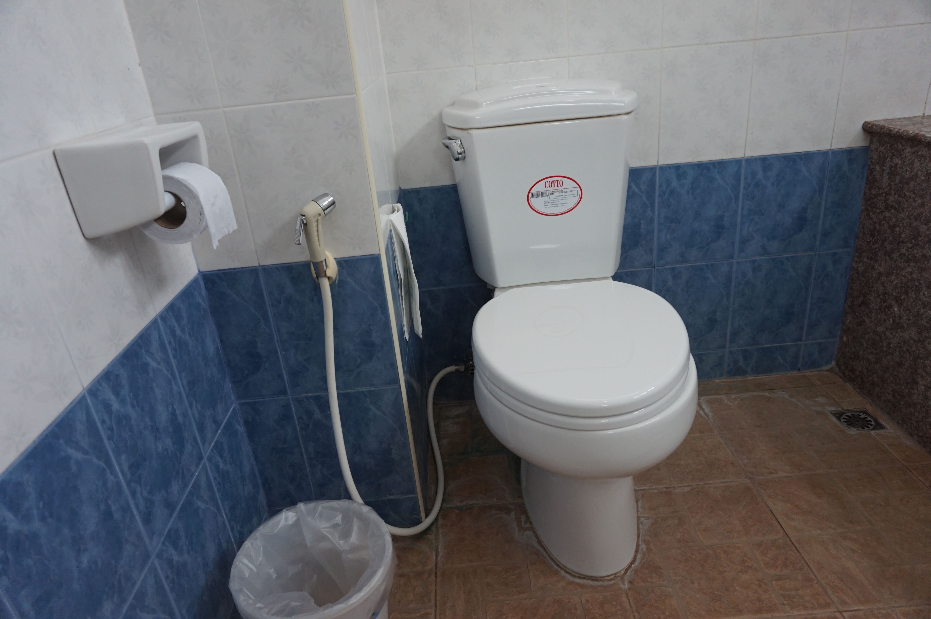 Un baño con un inodoro, un dispensador de toallas de papel, un cubo de basura de plástico y una manguera de ducha.
