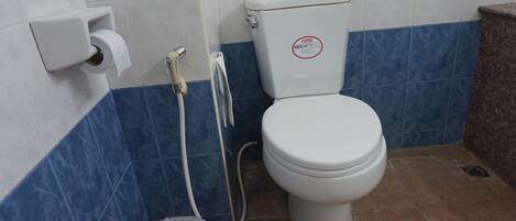 Un baño con un inodoro, un dispensador de toallas de papel, un cubo de basura de plástico y una manguera de ducha.