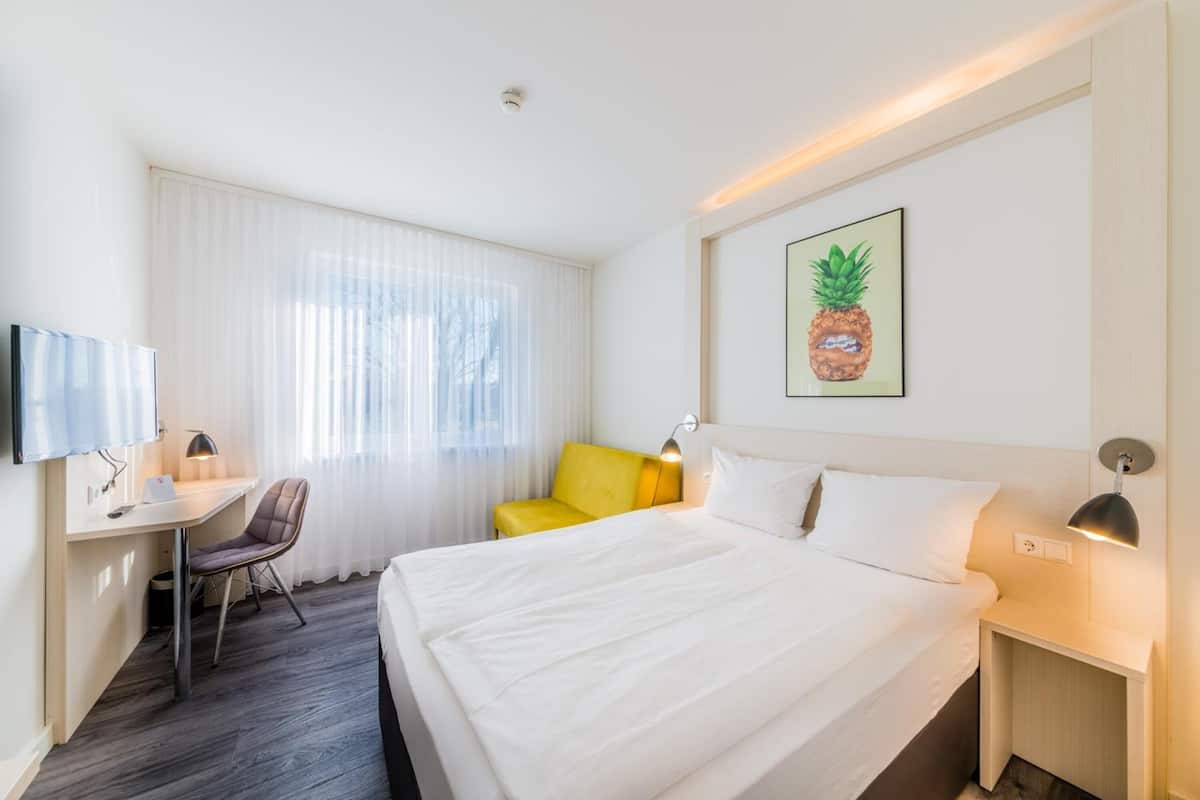 Habitación de hotel con cama, escritorio con silla, televisor y un cuadro de una piña.