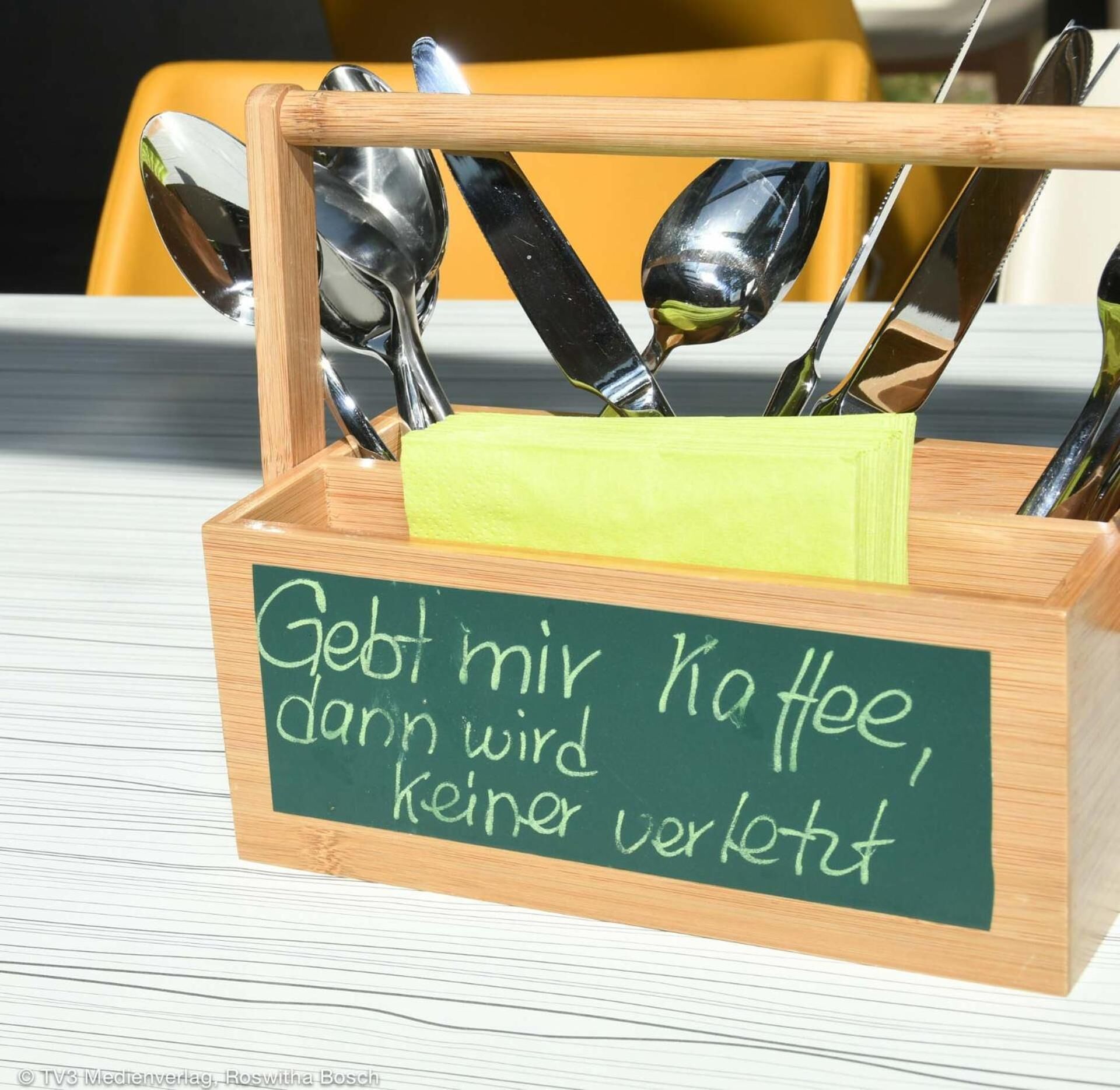 A wooden box with a chalkboard sign that reads "Gebt mir Kaffee, dann wird keine Gewalt" and contains several spoons.