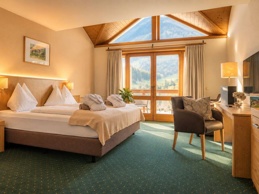 A hotel room with a bed, a desk, a chair, a TV, and a large window with a mountain view.