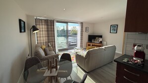 A modern living room with a sofa, a glass dining table, a TV, and a kitchen area with cabinets and a red appliance. - Clyde Waterfront Apartments (Glasgow)