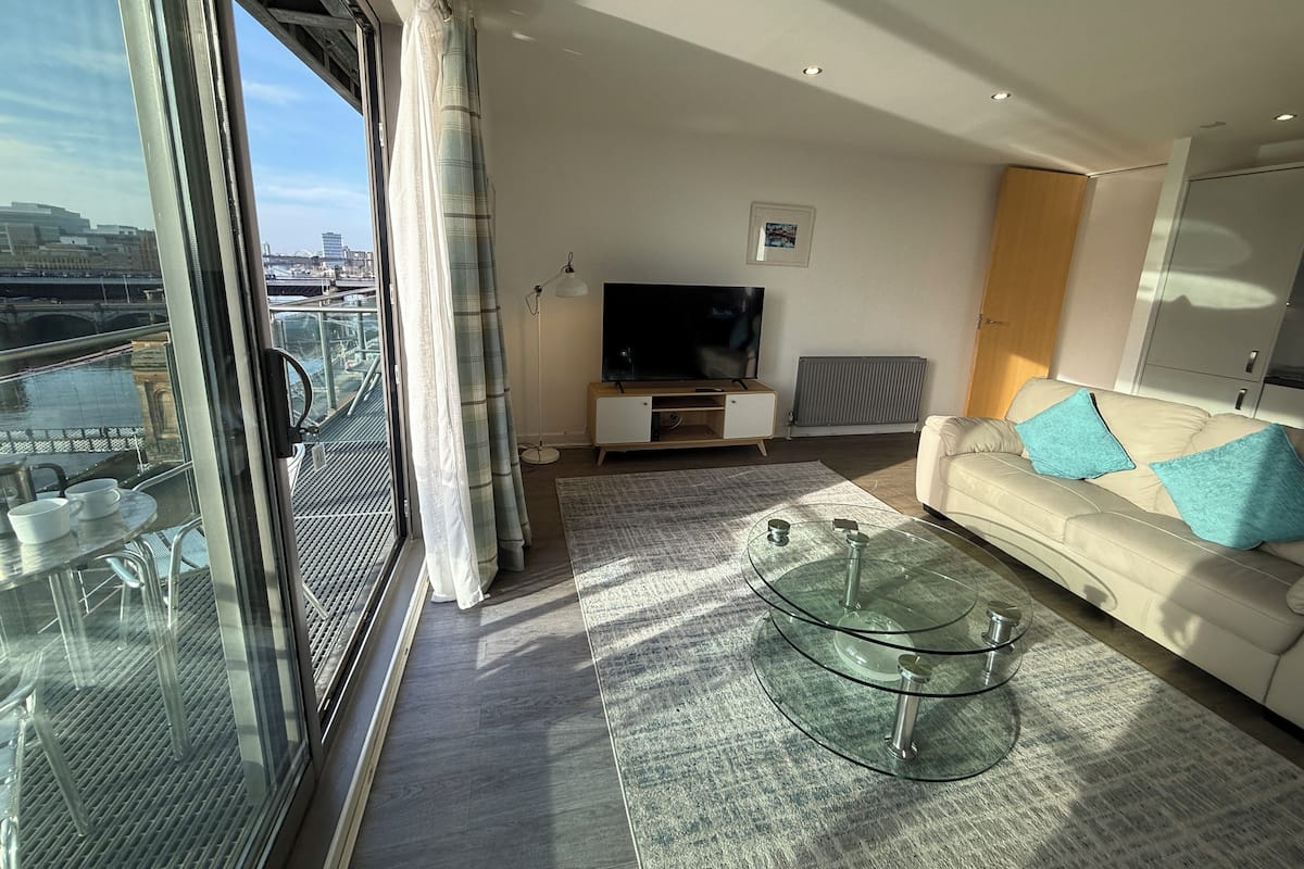 Un salon moderne avec une table basse en verre, un canapé avec des coussins bleus, un téléviseur à écran plat et une vue sur un pont et le paysage urbain, le tout visible à travers de grandes fenêtres.