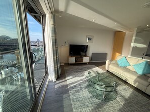 Ein modernes Wohnzimmer mit einem Glastisch, einer blauen Sofaausstattung, einem Flachbildfernseher und Blick auf eine Brücke sowie die Skyline durch große Fenster.