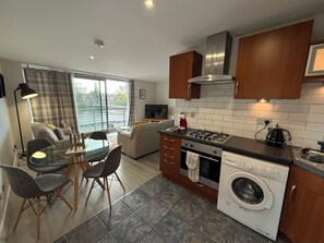 A modern kitchen with a stove, oven, and washing machine, a dining area with chairs and a table, and a sofa. - Clyde Waterfront Apartments (Glasgow)