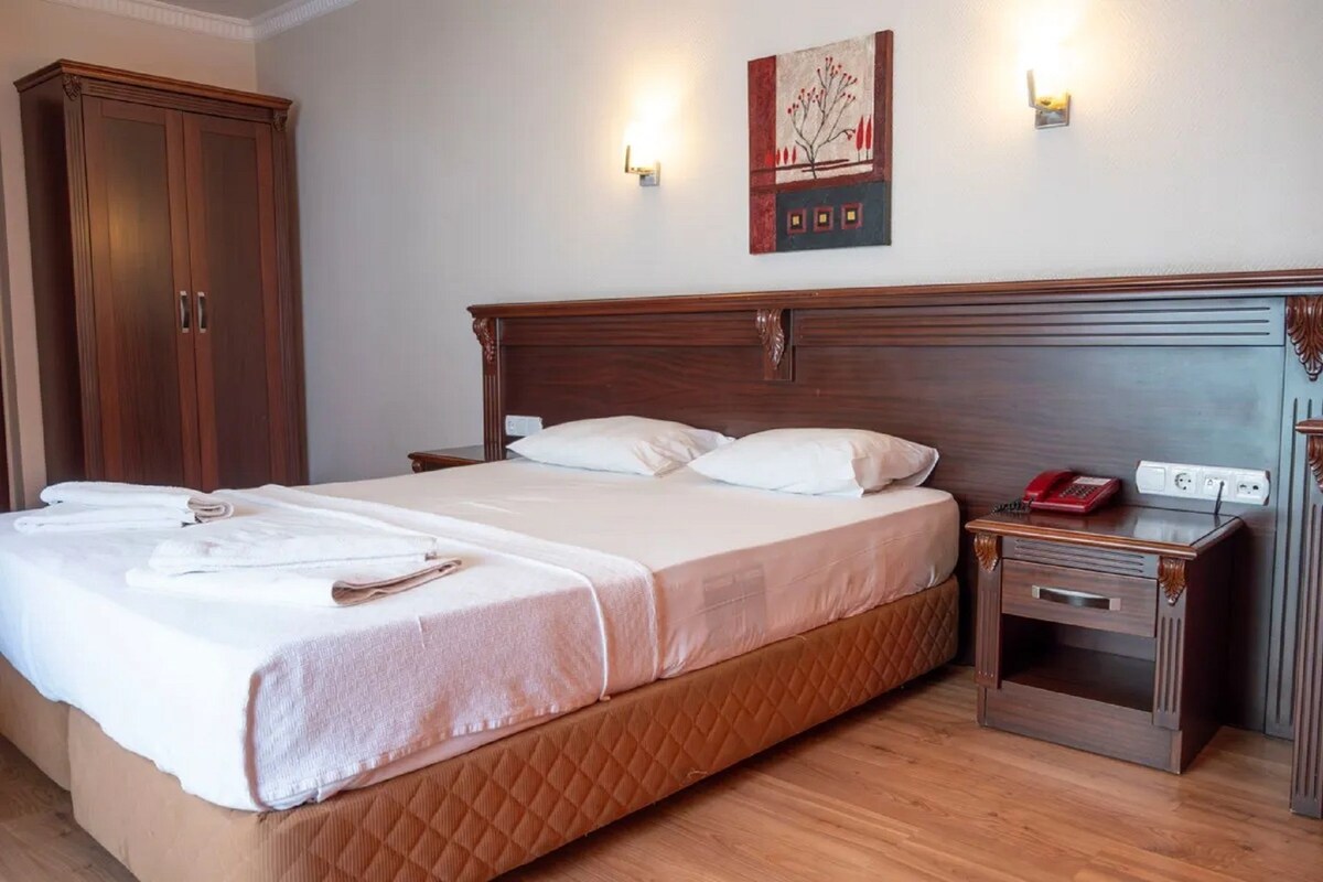 A neatly made bed with a quilted cover, a wooden nightstand with a telephone, and a wall-mounted light fixture.