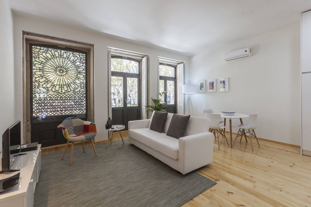 A modern living room with a white sofa, a small round table, and a colorful chair. There are two large windows with decorative glass panels, a potted plant, and a flat-screen TV on a stand.