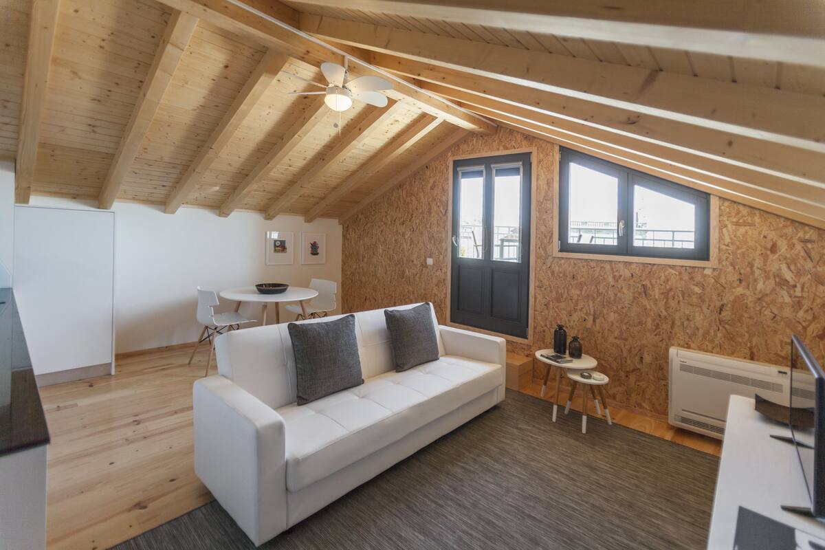 A cozy loft-style room with a white sofa, a small round table, a ceiling fan, and a large window offering a view of the outside.