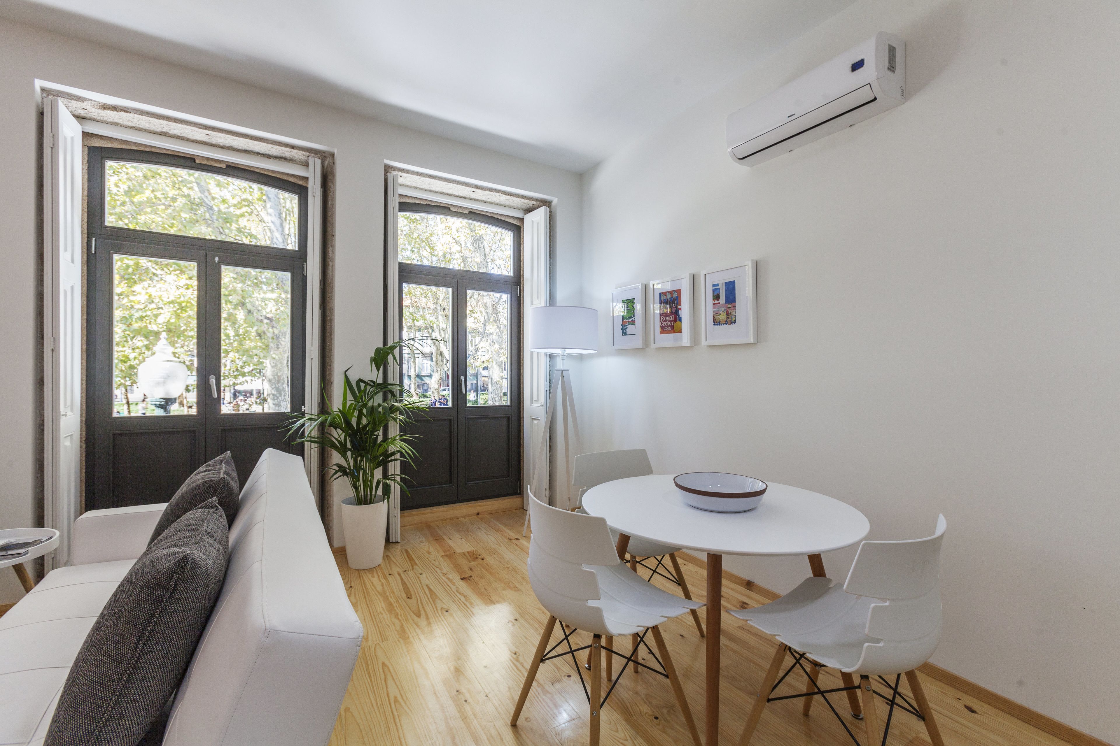 A modern living room with a white sofa, a dining table with chairs, a potted plant, and large windows with a view of trees.