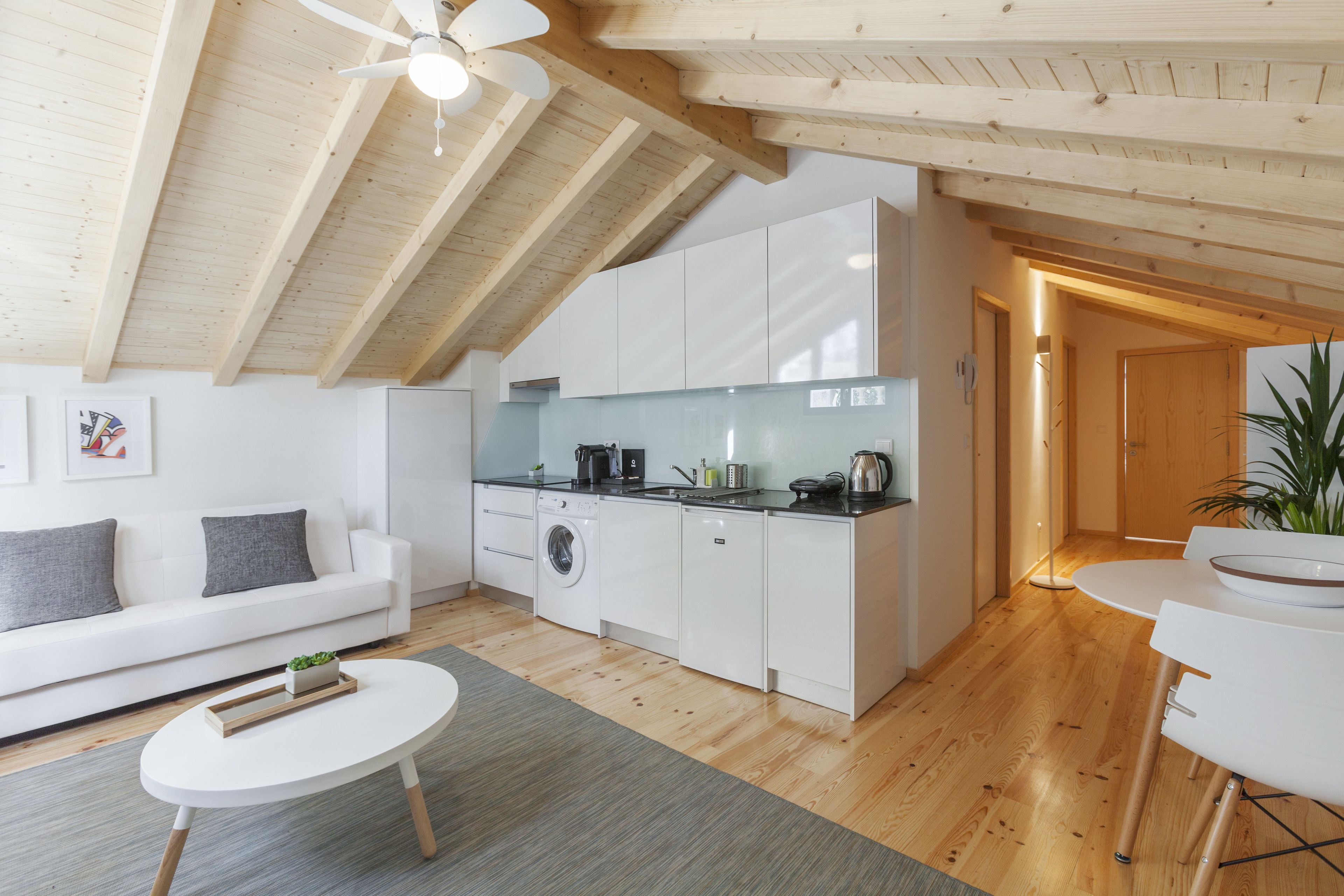 A modern kitchen with white cabinets, a built-in oven, and a coffee machine. There is a wooden ceiling with exposed beams and a ceiling fan.