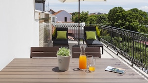 A wooden table with a potted plant, a bottle of orange juice, and a magazine on a balcony with two lounge chairs and a large umbrella. - BO - Marquês Apartments (Porto)