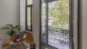A room with a large sliding glass door leading to a balcony with a wrought iron railing. There is a potted plant and a chair with a colorful cushion. - BO - Marquês Apartments (Porto)