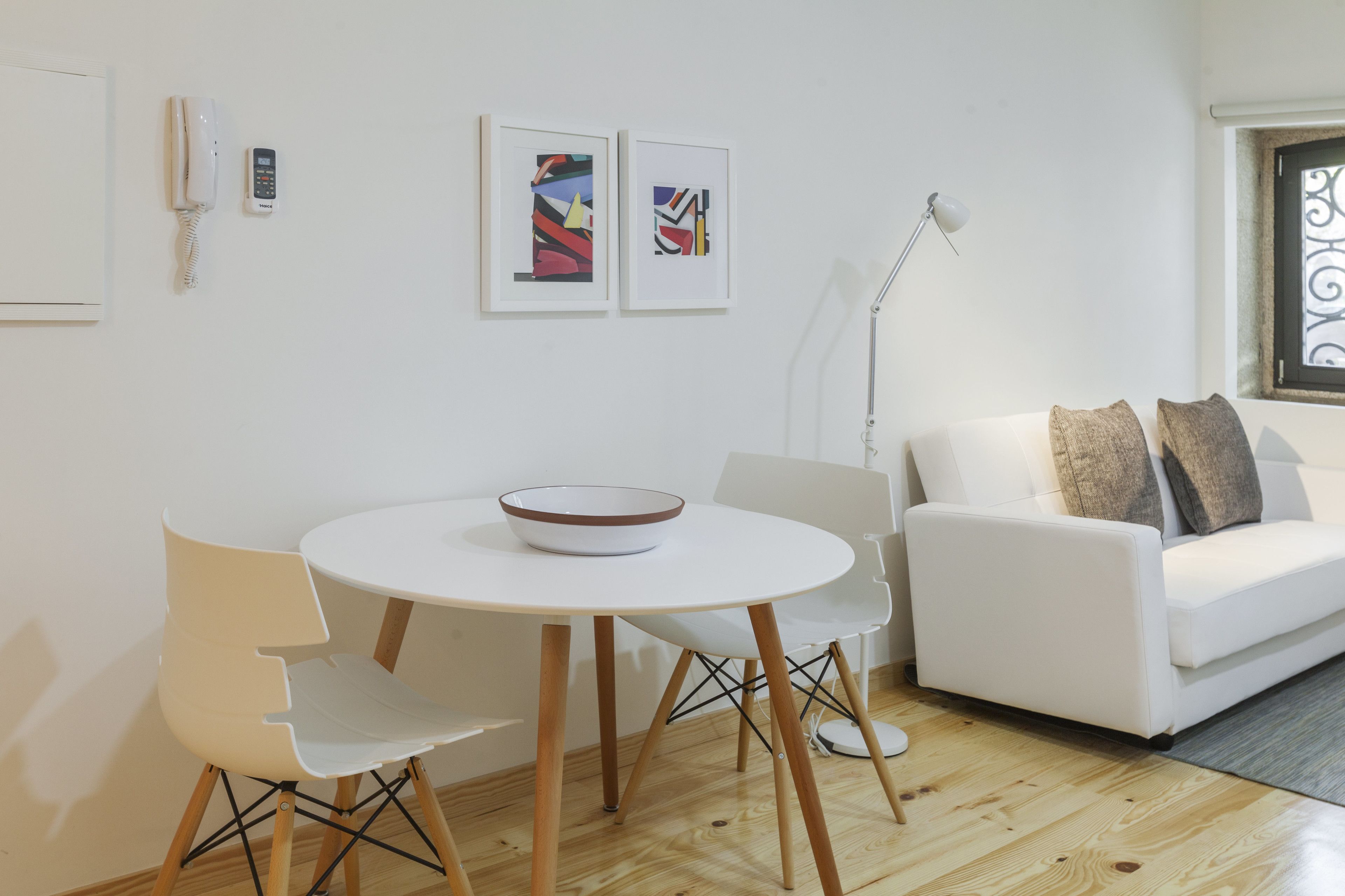 A modern living room with a white sofa, a round dining table with a bowl, and two chairs. There are two framed artworks on the wall, a white telephone, and a floor lamp.