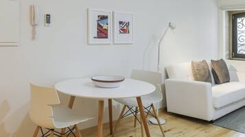 A modern living room with a white sofa, a round dining table with a bowl, and two chairs. There are two framed artworks on the wall, a white telephone, and a floor lamp.