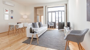 A modern living room with a white sofa, a dining table with chairs, a patterned armchair, and a large window with a view. - BO - Marquês Apartments (Porto)