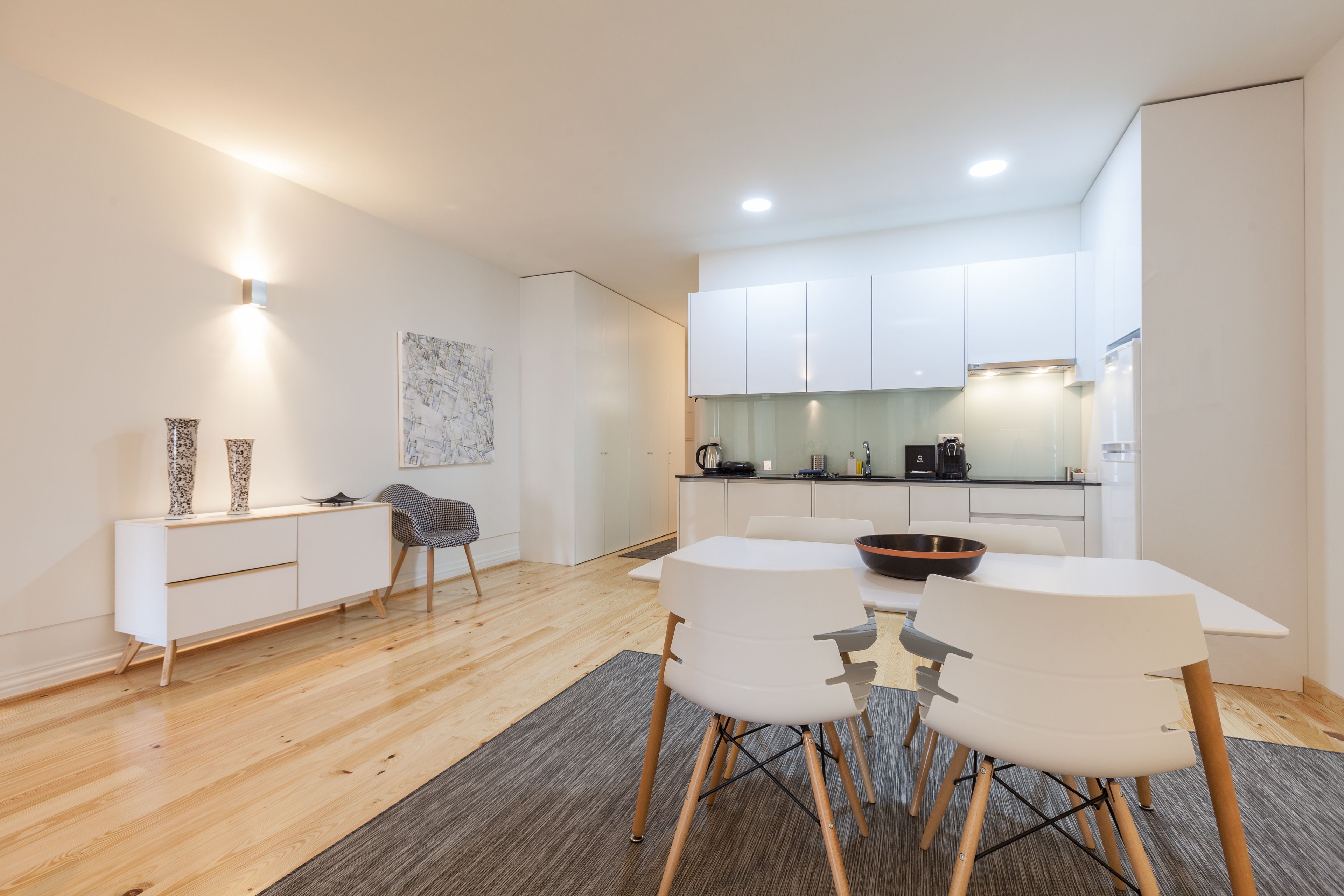 Een moderne keuken met witte kastjes, een eethoek met een witte tafel en stoelen, en een grijze vloerkleed.