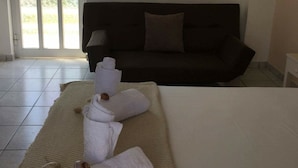 Une chambre d’hôtel avec un canapé de couleur sombre, un téléviseur à écran plat fixé au mur et un grand lit avec du linge de lit blanc et un plaid beige.