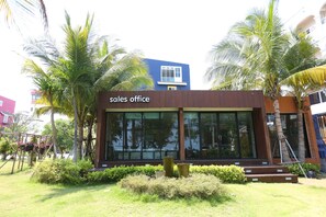 A modern sales office with large windows, surrounded by well-maintained greenery and palm trees.