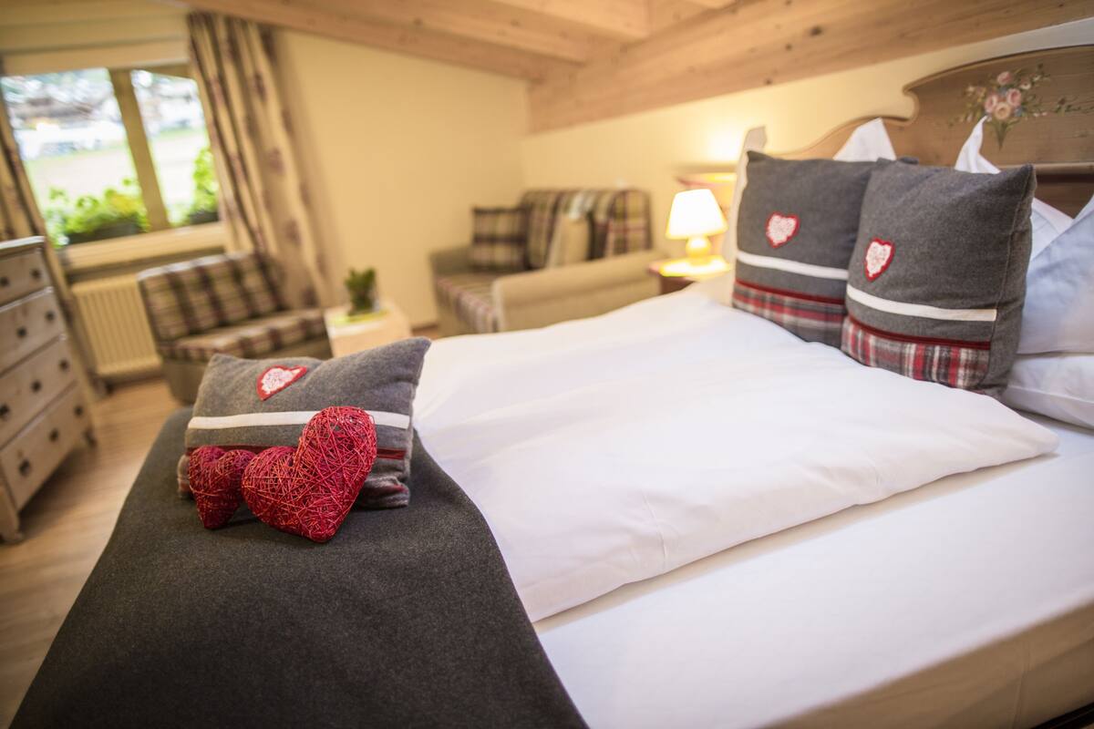 Una cama bien hecha con edredón gris y blanco, dos almohadas con diseños de corazones y una almohada con forma de corazón roja.