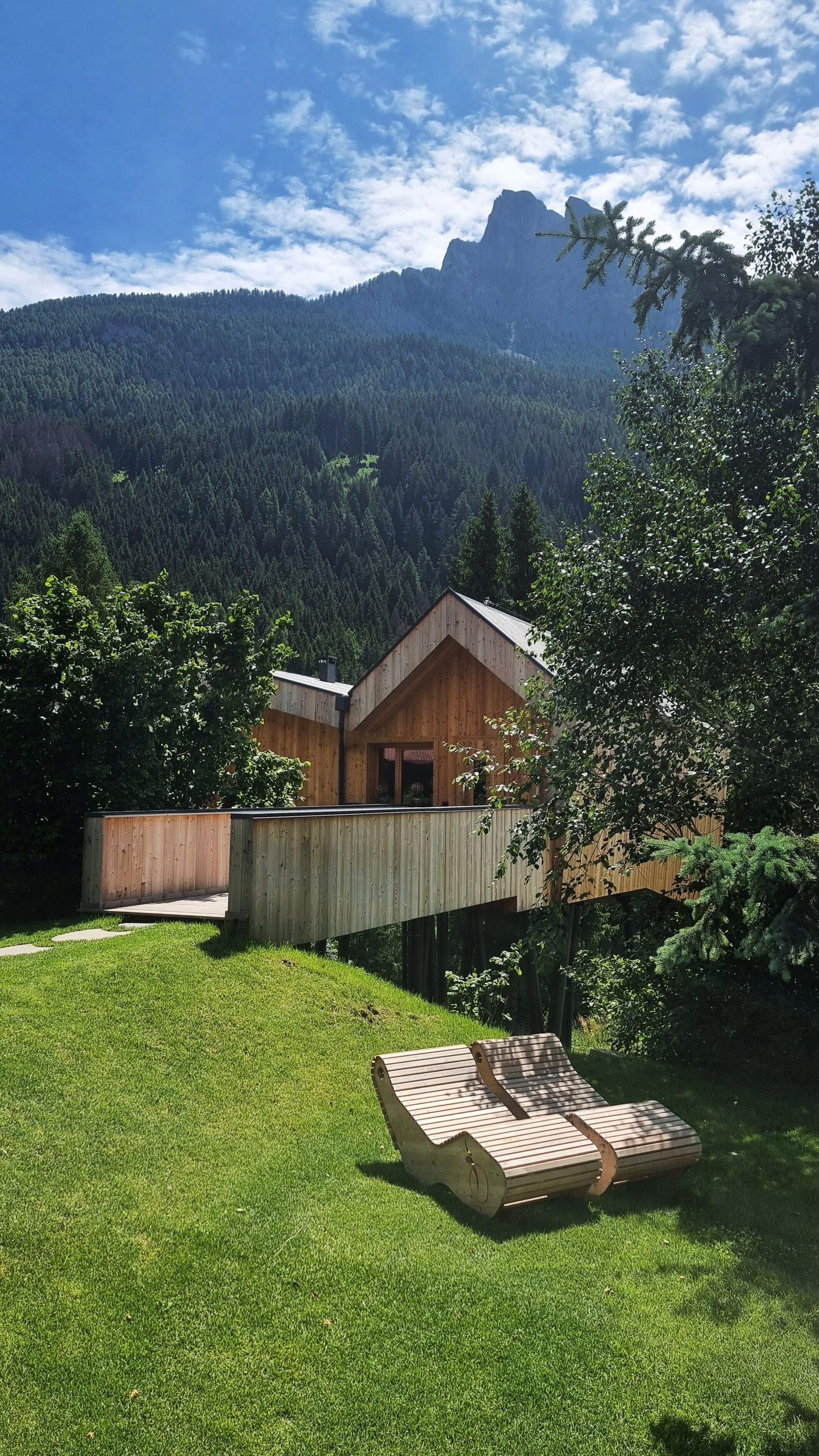 A wooden cabin with a deck, surrounded by a lush green lawn and trees, with a mountainous backdrop.
