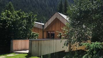 A wooden cabin with a deck, surrounded by a lush green lawn and trees, with a mountainous backdrop.