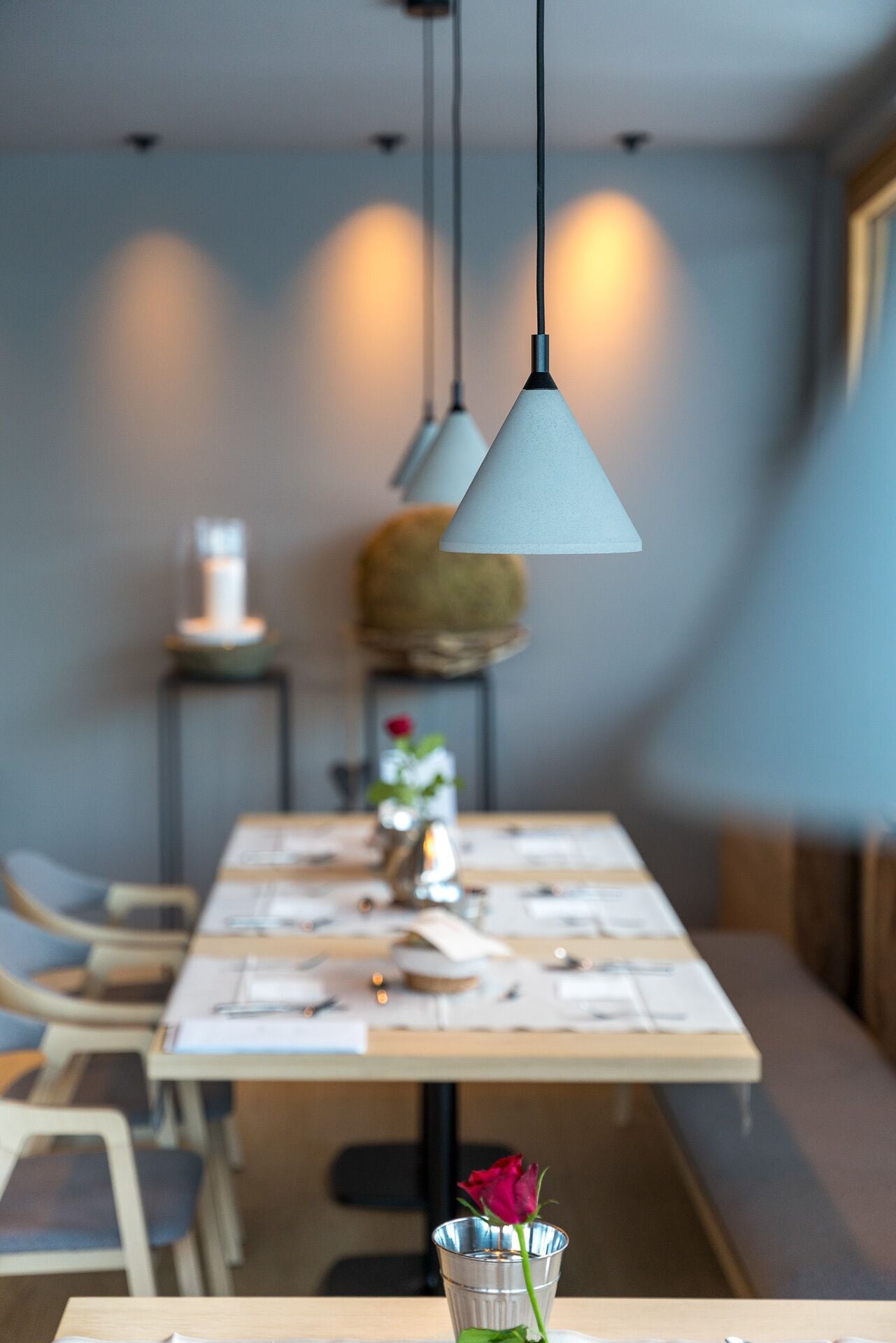 A dining area with a table set for two, a vase with a single rose, and pendant lights above.