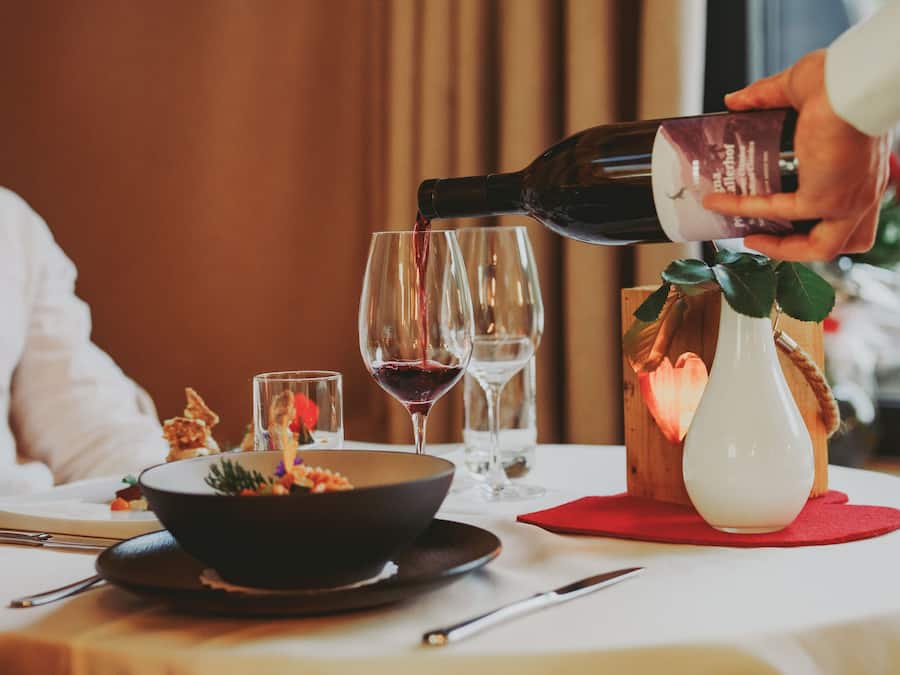 Mesa de jantar com um prato de comida, copos de vinho e uma garrafa sendo servida em um dos copos.