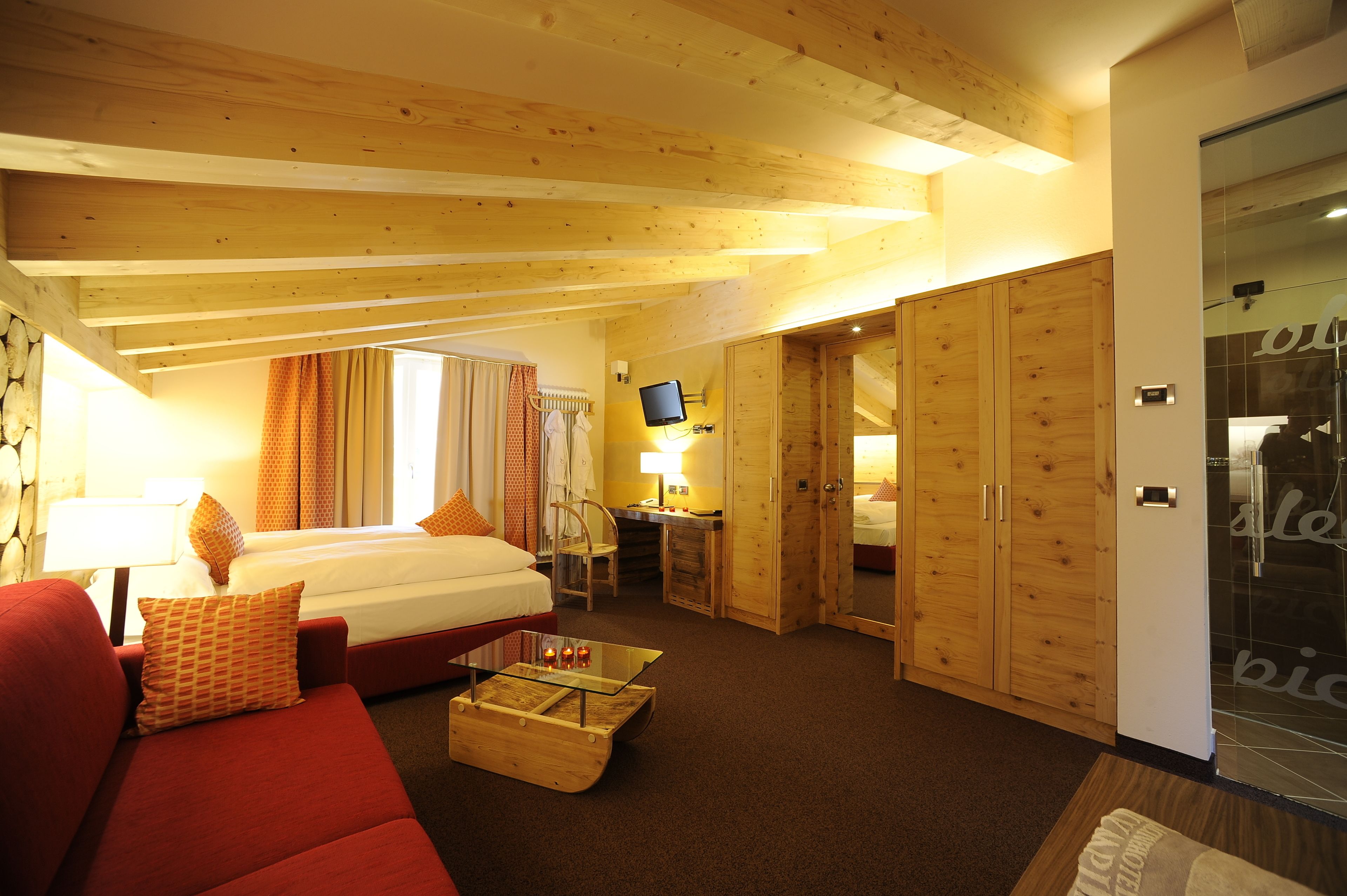 Habitación de hotel con cama, sofá rojo, mesa de centro de vidrio, escritorio con computadora y armarios de madera.