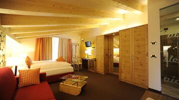 Habitación de hotel con cama, sofá rojo, mesa de centro de vidrio, escritorio con computadora y armarios de madera.