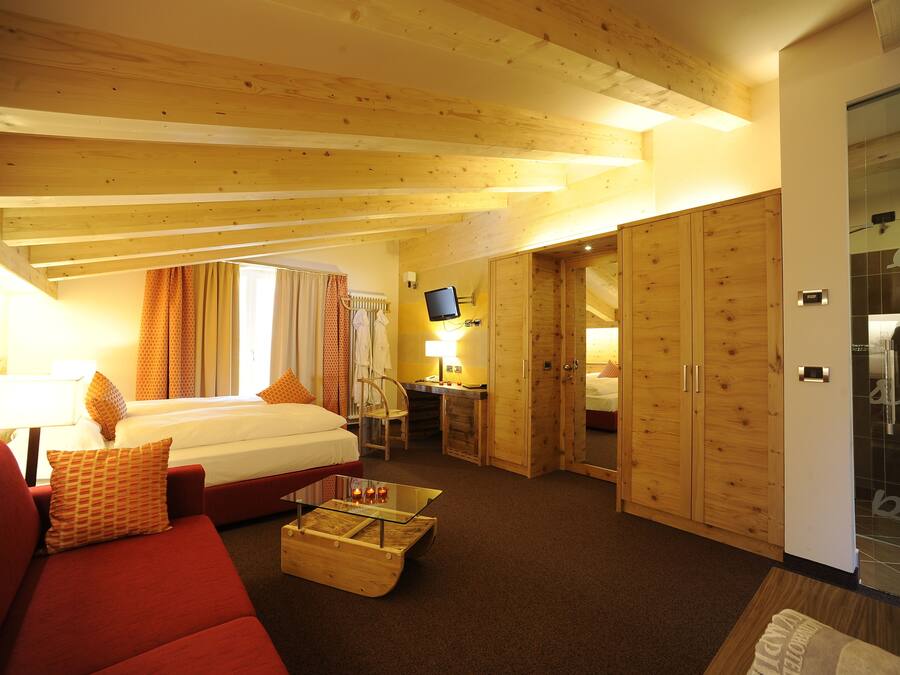 Habitación de hotel con cama, sofá rojo, mesa de centro de vidrio, escritorio con computadora y armarios de madera.