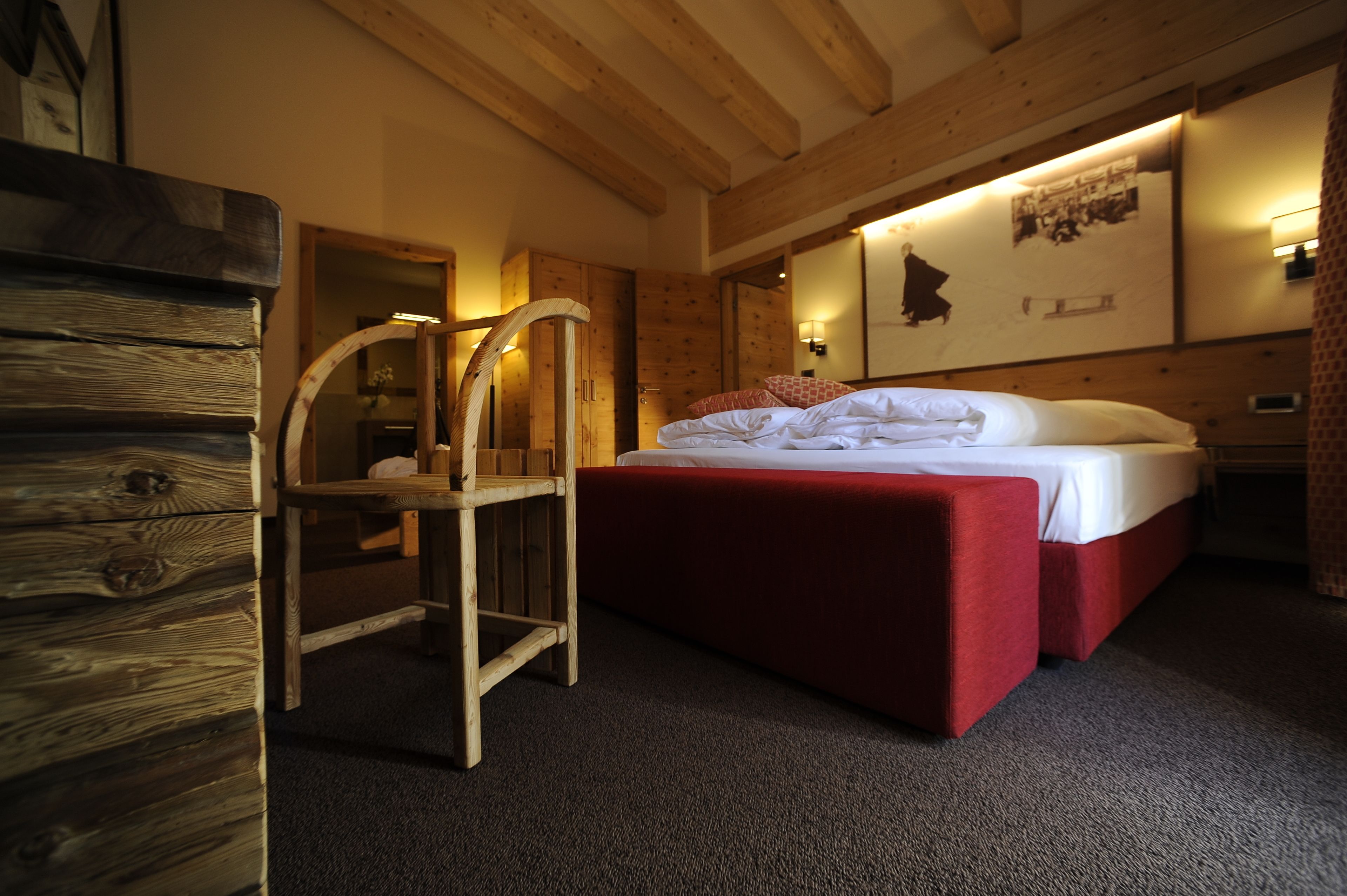A bedroom with a wooden bed frame, a red upholstered bench, a wooden chair, and a mirror.