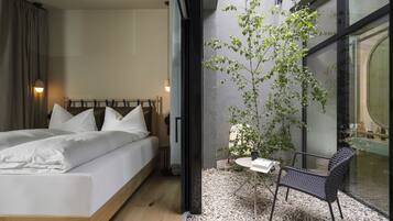 A modern bedroom with a bed, a small table, and a chair. There is a window with a view of a tree and a building.