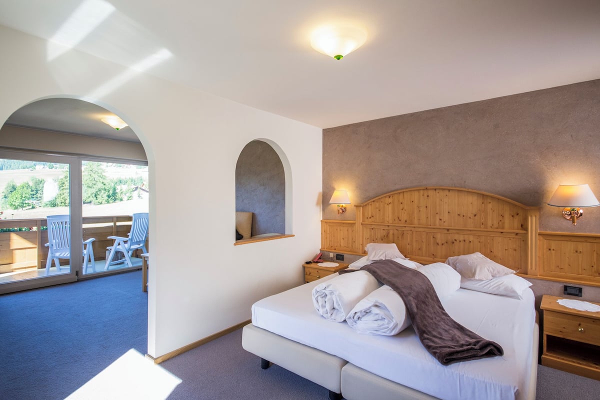 Habitación de hotel con una cama grande, cabecera de madera, dos mesitas de noche y vistas al balcón.