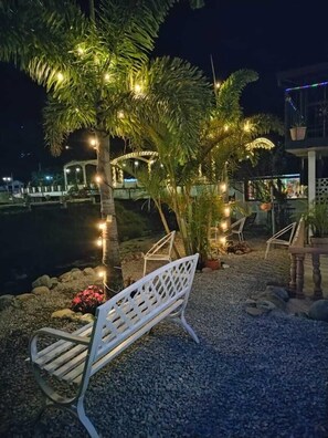 Un espacio de estar al aire libre con sillas y banca de metal blanco, rodeado de palmeras y luces decorativas.