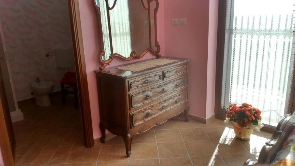 Una habitación con un aparador de madera, un espejo, una ventana con persianas y un florero con flores.