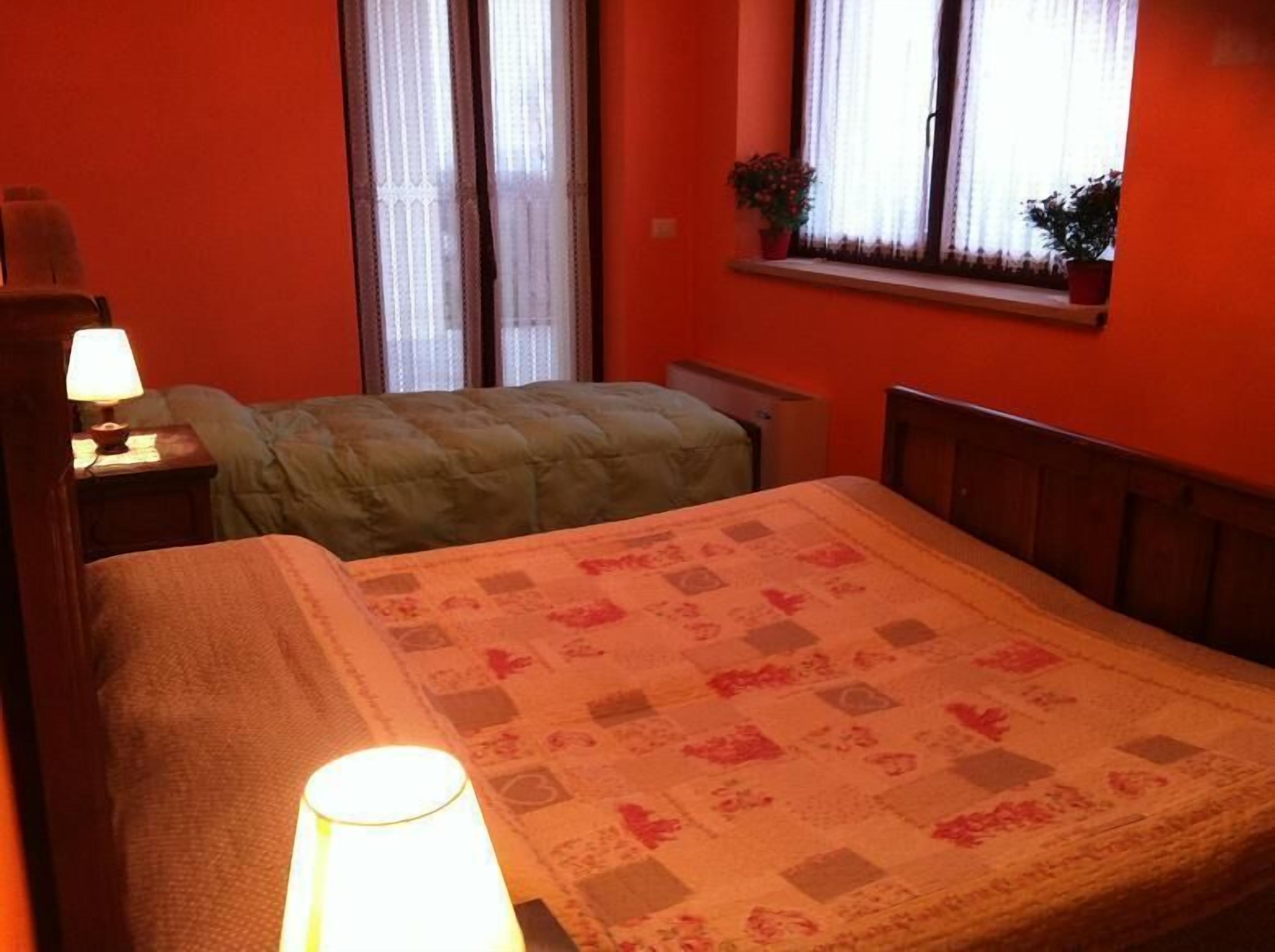 Ein Zimmer mit zwei Betten, einem Fenster mit Vorhängen, einem Nachttisch mit Lampe und einem Blumentopf auf der Fensterbank.