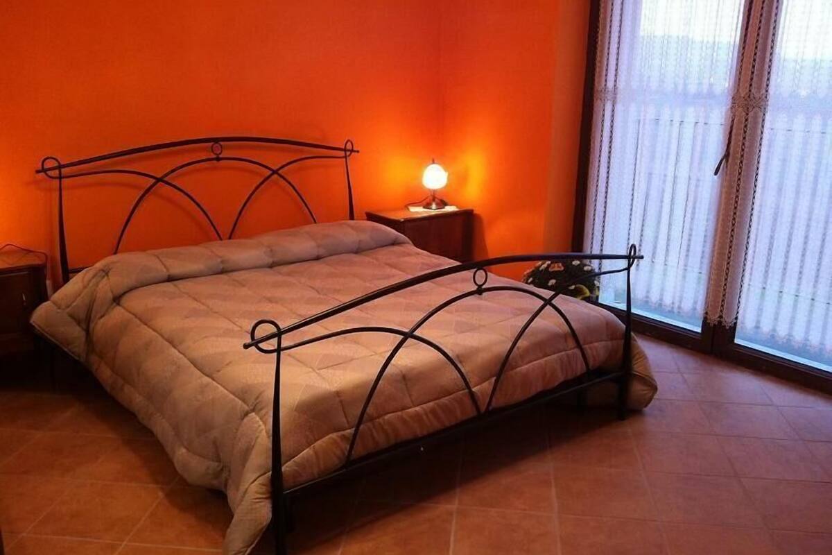 A bed with a metal frame and a beige quilt in a room with orange walls and a window with blinds.