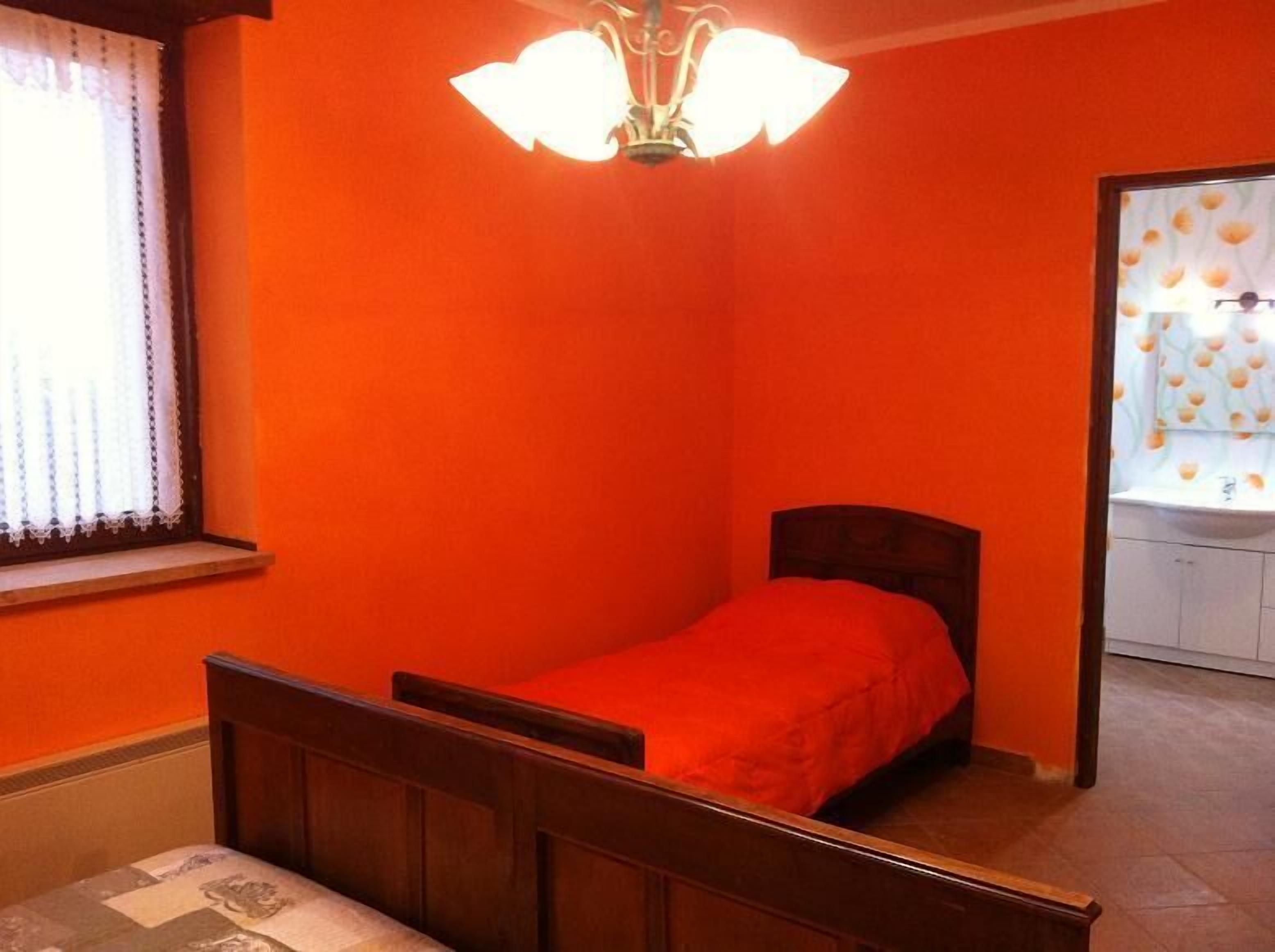 A small, single-bed room with a wooden headboard, a red bedspread, and a white ceiling light fixture.