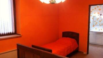 A small, single-bed room with a wooden headboard, a red bedspread, and a white ceiling light fixture.