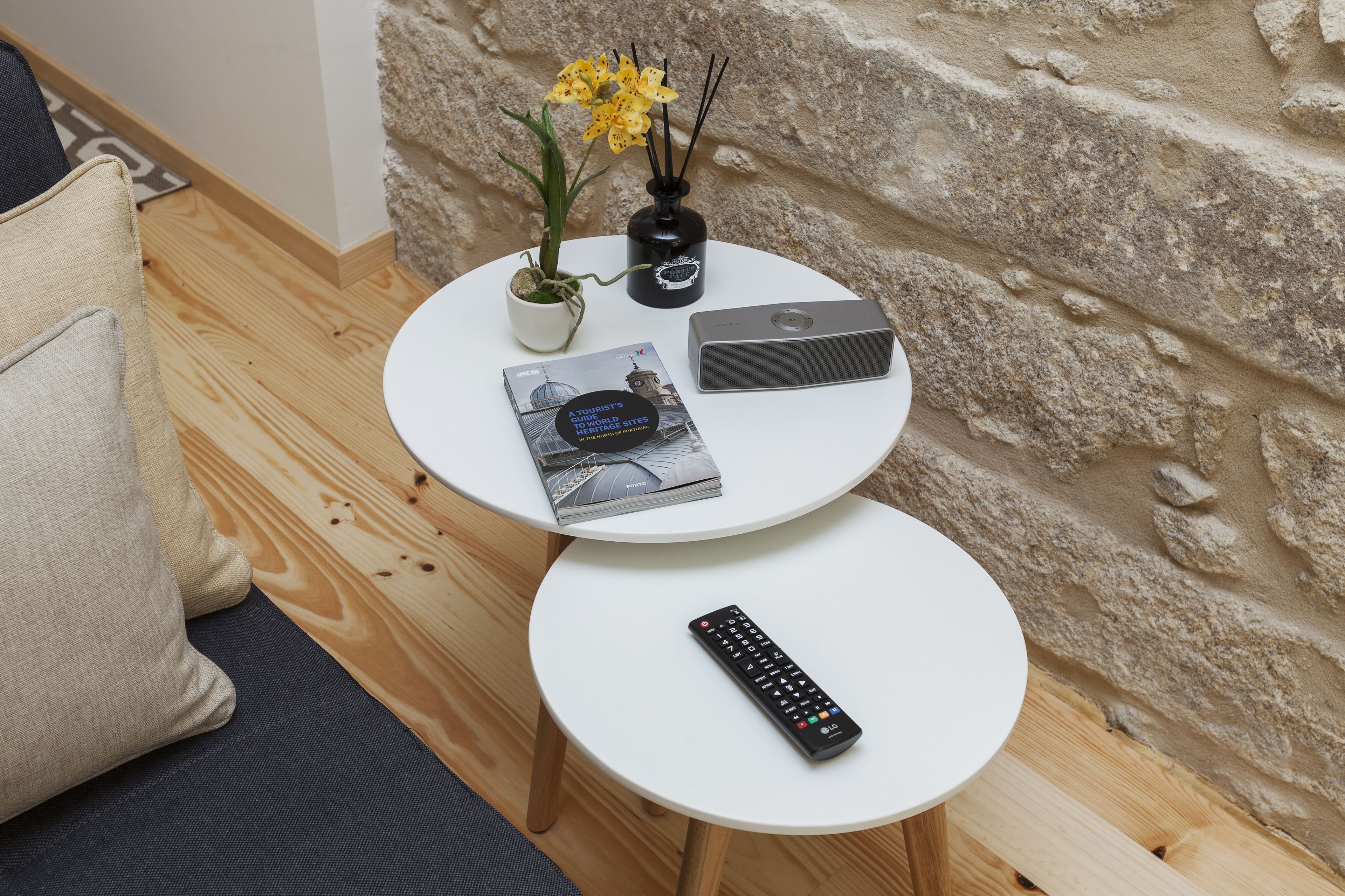 Una mesa redonda pequeña con un control remoto, un libro, un jarrón decorativo con flores y un altavoz.