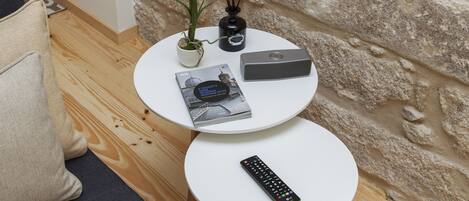 Una mesa redonda pequeña con un control remoto, un libro, un jarrón decorativo con flores y un altavoz.