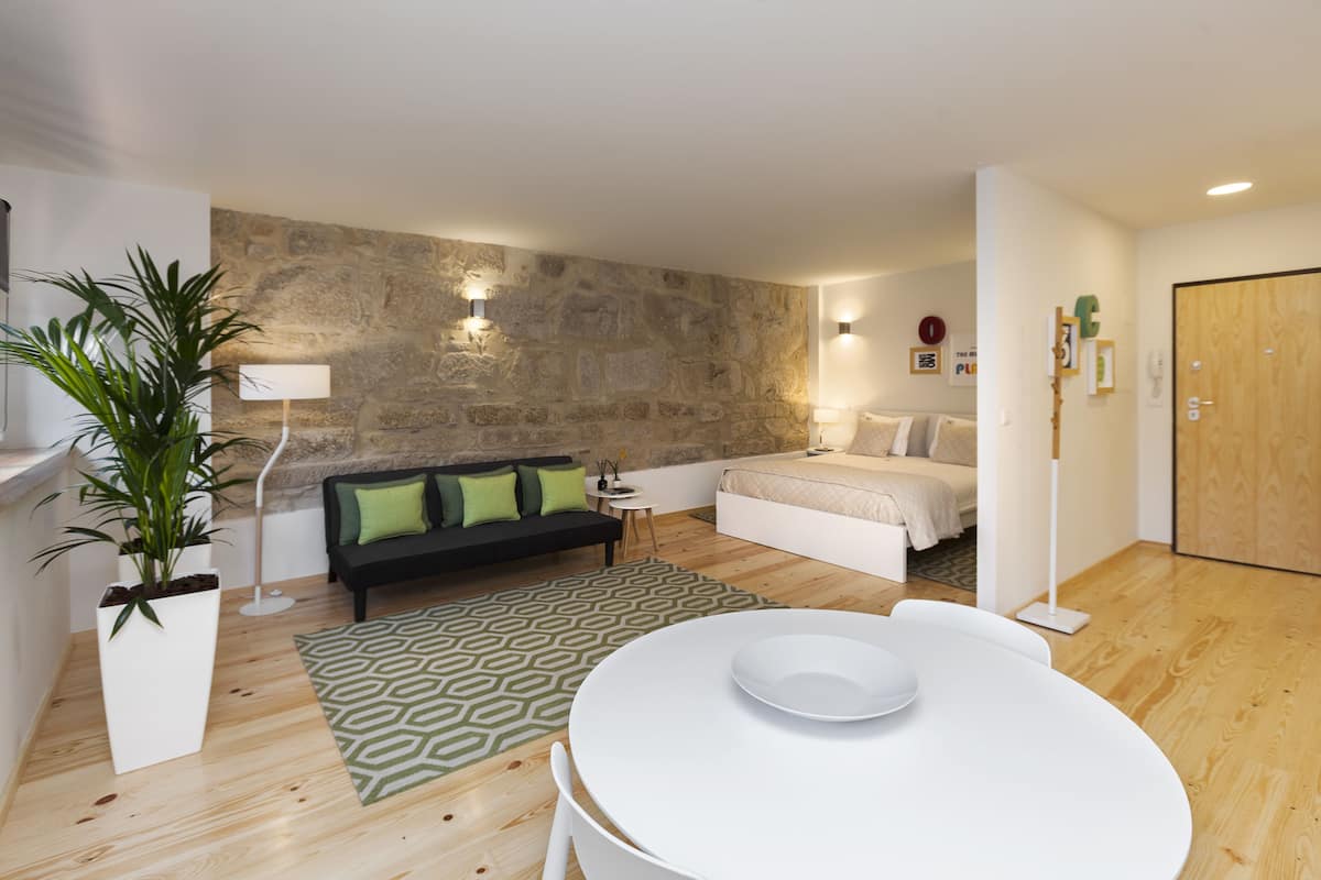 Un salon moderne avec un mur d’accent en pierre, un canapé noir avec des coussins verts, une table ronde blanche et un lit en arrière-plan.
