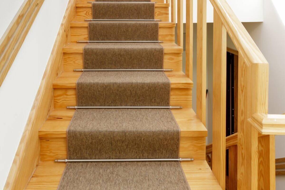 Un escalier en bois, dont les marches sont recouvertes de moquette, mène à un palier où se trouve une porte blanche et une fenêtre au-dessus.