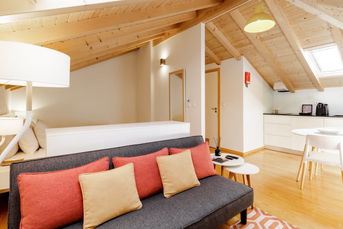 Un appartement moderne de style « loft » avec un lit en mezzanine, un canapé gris agrémenté de coussins rouges et beige, un coin repas avec une table et des chaises blanches, et des poutres en bois au plafond.