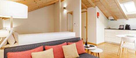 A modern loft apartment with a loft bed, a gray sofa with red and beige cushions, a dining area with a white table and chairs, and wooden beams on the ceiling.