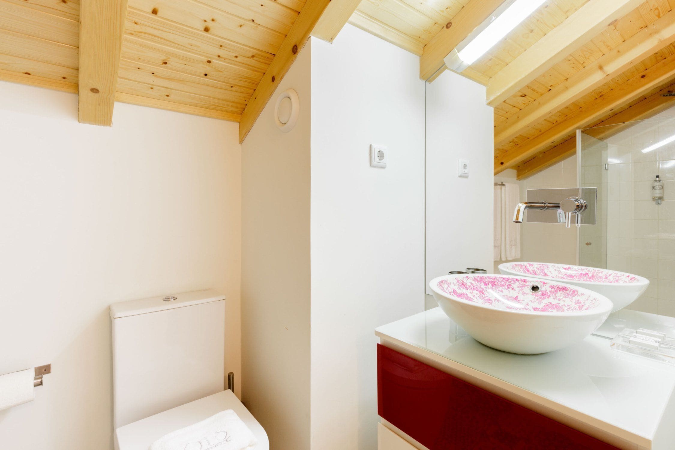 Un baño con vigas de madera, paredes blancas, una encimera roja y dos lavamanos blancos con motivos florales rosados.