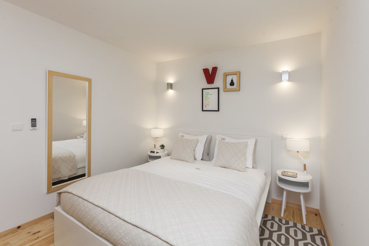 Une chambre à coucher bien rangée, avec un grand lit, des tables de chevet avec des lampes, un miroir fixé au mur et un cadre contenant une citation inspirante.