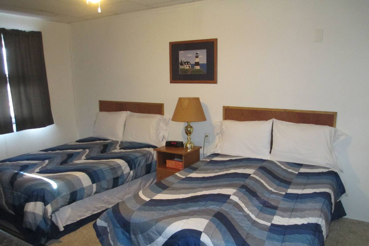 Ein Hotelzimmer mit zwei Betten, einem Nachttisch mit Lampe, einem gerahmten Bild an der Wand und einem Fenster mit Vorhängen.