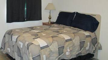 Una cama con una colcha estampada, una lámpara de noche y un almohadón oscuro.
