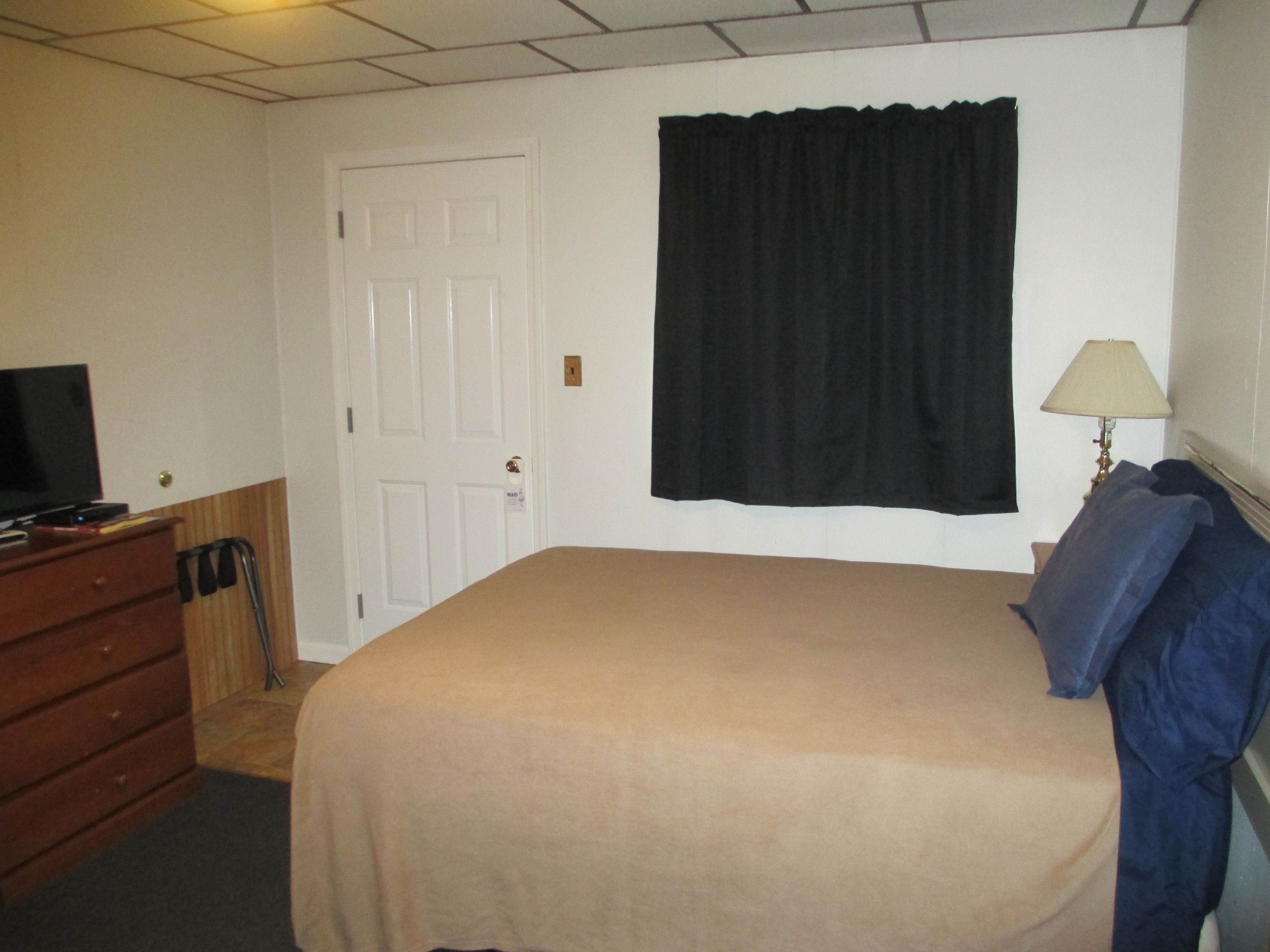 A bedroom with a bed, a dresser, a television, a door, and a window with a curtain.